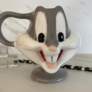 May include: A ceramic mug shaped like Bugs Bunny, with a gray and white color scheme. The mug has a handle and a base.