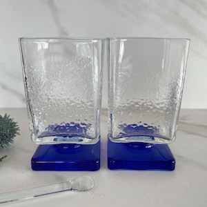 May include: Two clear glass tumblers with a textured surface and a blue square base. The glasses are empty and sitting on a white surface.