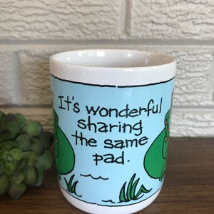 May include: White ceramic mug with a blue background and two green frogs. The text on the mug reads "It's wonderful sharing the same pad."