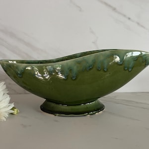 May include: An elongated, boat-shaped ceramic bowl with a glossy olive green glaze. The rim features a decorative drip pattern in a lighter, muted teal tone. The vessel is supported by a circular pedestal base and has a smooth, curved silhouette.