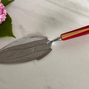 May include: A silver-toned metal cake server with a pointed, leaf-shaped blade featuring a decorative zigzag engraved border. The handle is composed of a vibrant red and gold-toned striped design, connecting to the blade with a polished silver ferrule.