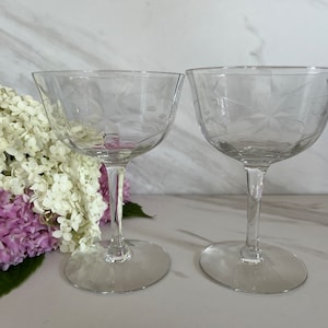 May include: Two clear, stemmed champagne glasses with etched floral designs. The glasses have a wide, shallow bowl and a long, slender stem. The base is round. The glasses are set against a backdrop of white and purple flowers.