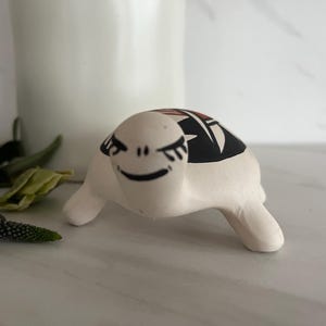 May include: A ceramic turtle figurine with a white body and a black and red patterned shell. The turtle has a smiling face with black details. The figurine is on a white surface with purple flowers and a white candle in the background.