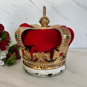 May include: A gold-colored crown with red velvet interior, adorned with colorful jewels. The crown has a white base with text. A decorative item, possibly for display or costume use.