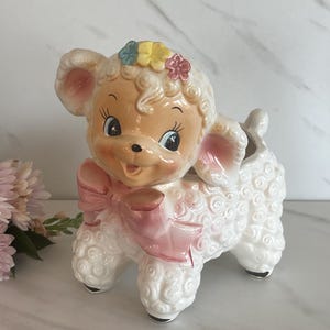 May include: A vintage ceramic lamb planter with a smiling face, adorned with a pink bow and a floral crown. The lamb has white, textured fur and black hooves. The planter is a charming decorative piece.