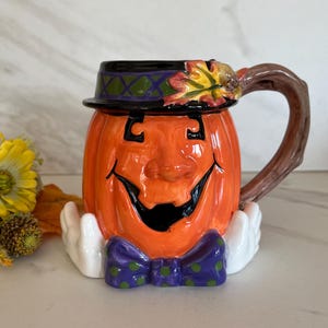 May include: Orange ceramic Halloween mug shaped like a jack-o'-lantern with a black hat, purple bow tie, and white gloves. The handle is brown and shaped like a tree branch. The mug has a smiling face with black eyes and mouth.