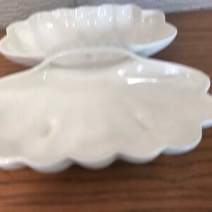 May include: Two white, shell-shaped ceramic dishes. The dishes have a scalloped edge and a smooth, glossy finish. The dishes are empty and appear to be new. They are sitting on a wooden surface.