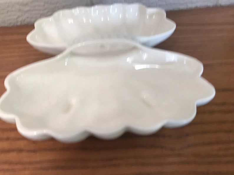 May include: Two white, shell-shaped ceramic dishes. The dishes have a scalloped edge and a smooth, glossy finish. The dishes are empty and appear to be new. They are sitting on a wooden surface.