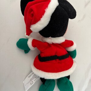 Puede incluir: Un Mickey Mouse de peluche vestido con un traje de Pap&aacute; Noel rojo con ribete de piel blanca, guantes verdes y zapatos verdes. El peluche tiene una etiqueta negra con el texto "Disney Store" y "CE".