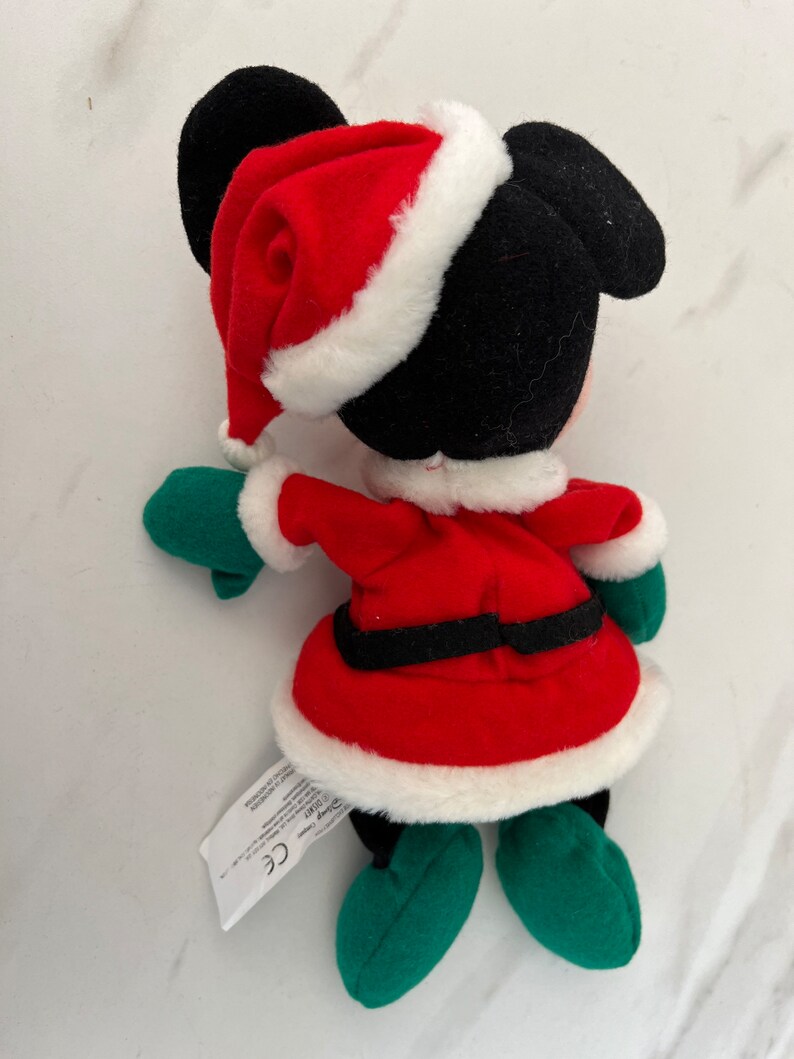 Puede incluir: Un Mickey Mouse de peluche vestido con un traje de Pap&aacute; Noel rojo con ribete de piel blanca, guantes verdes y zapatos verdes. El peluche tiene una etiqueta negra con el texto "Disney Store" y "CE".