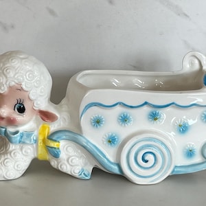 May include: A white ceramic planter shaped like a lamb pulling a cart. The lamb has a blue bow and yellow ribbon. The cart features blue scalloped trim, blue floral accents, and a spiral wheel design. The finish is glossy with painted details.