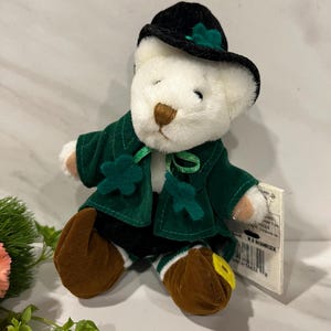 May include: A white teddy bear wearing a green velvet jacket, black hat, and brown shoes. The hat and jacket are adorned with shamrock accents. The bear is a St. Patrick's Day themed plush toy.