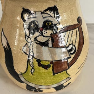May include: A beige ceramic pitcher featuring a hand-painted illustration of a cat with black and white fur, wearing a green dress and a beaded necklace. The cat is depicted playing a brown harp, with a long black tail extending along the side.