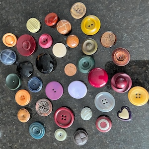 May include: A collection of various buttons in different colors, shapes, and sizes. The buttons include round, oval, and heart-shaped designs, with colors ranging from yellow and orange to blue and black. Some buttons have multiple holes.