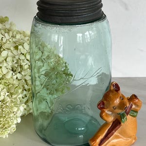 May include: A vintage blue-green Mason jar with a dark gray lid. A small, orange ceramic deer figurine with a green scarf and a brown stick is next to the jar. Dried white flowers are in the background.