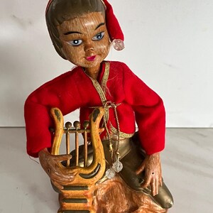 May include: A figurine of a figure with a wood-grain finish wearing a red cap and red jacket with gold trim, holding a small yellow lyre. The figure has blue eyes, pink lips, and metallic gold-colored pants, seated on a textured base.