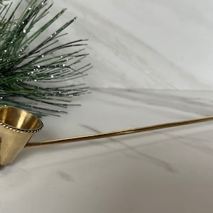 May include: A gold candle snuffer with a decorative, scalloped design and a round, brown knob on the end of the handle.