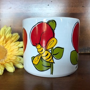 May include: White ceramic mug with a red, yellow and green floral design featuring a bee. The flowers are stylised and have a red centre.