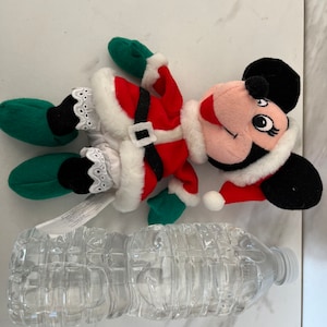 Puede incluir: Un juguete de peluche de Minnie Mouse vestida de Pap&aacute; Noel rojo y blanco. El juguete lleva un gorro verde y zapatos verdes. El juguete est&aacute; tumbado sobre una superficie blanca.