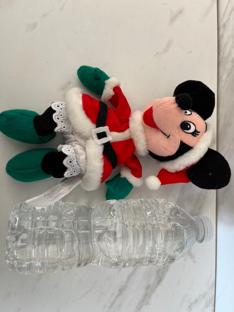 Puede incluir: Un juguete de peluche de Minnie Mouse vestida de Pap&aacute; Noel rojo y blanco. El juguete lleva un gorro verde y zapatos verdes. El juguete est&aacute; tumbado sobre una superficie blanca.