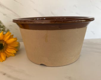 May include: A brown and tan ceramic pot with a wide rim. The top rim is a darker brown, and the body is a lighter tan color. A yellow sunflower is visible to the left of the pot.