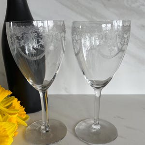 May include: Two clear glass wine glasses with etched floral designs around the top rim. The glasses are on a white surface.