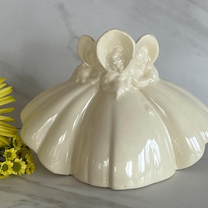 May include: Cream-colored ceramic figurine featuring three figures with wide-brimmed hats, atop a scalloped base. The figurine has a glossy finish and is placed next to a yellow flower.