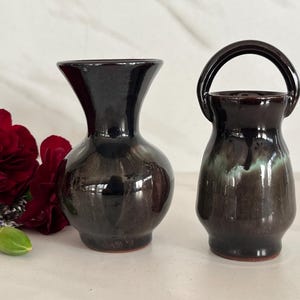 May include: Two ceramic vases with a glossy, dark brown glaze. One vase has a traditional shape, while the other has a handle. Red flowers are visible on the left side of the image. The vases are likely for decorative purposes.