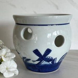 May include: A white ceramic bulb planter with a blue windmill design. The planter has two large circular openings and a blue band around the top. The base features a detailed illustration of a windmill and landscape.