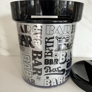 May include: A clear plastic ice bucket with a black lid and rim. The bucket has a silver metallic finish with the word "BAR" in various fonts and sizes printed on the exterior. The lid is partially removed.