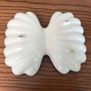 May include: A white, ceramic, bow-shaped dish with a glossy finish. The dish has a fluted design and is likely used for serving or display. The dish is on a wooden surface.