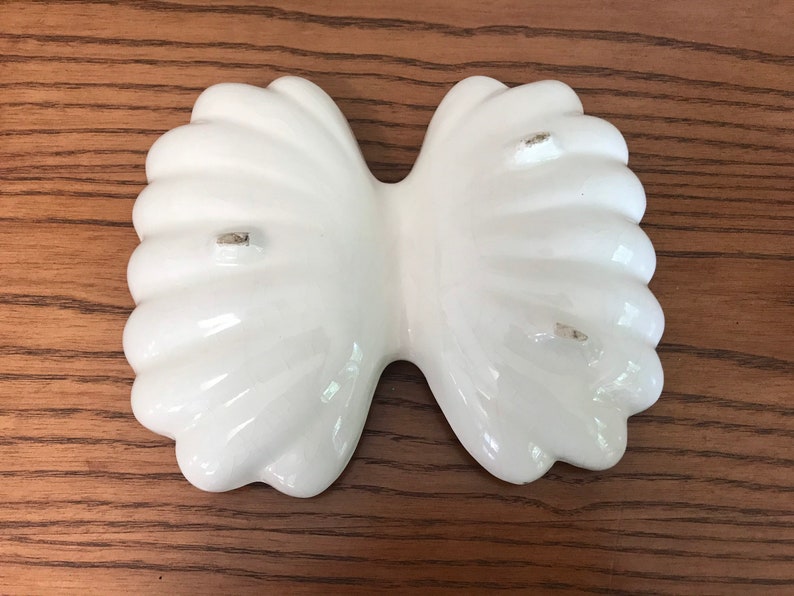 May include: A white, ceramic, bow-shaped dish with a glossy finish. The dish has a fluted design and is likely used for serving or display. The dish is on a wooden surface.