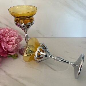May include: Two amber-colored glass candle holders with silver-colored metal bases. The candle holders have a decorative openwork design around the bowl. Pink peonies are in the background.