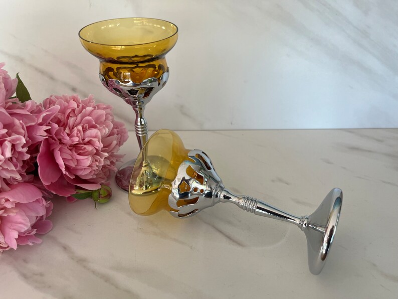 May include: Two amber-colored glass candle holders with silver-colored metal bases. The candle holders have a decorative openwork design around the bowl. Pink peonies are in the background.
