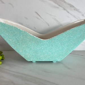 May include: A turquoise ceramic planter with a speckled texture and a V-shaped design. The interior is white, and the planter sits on small feet. A pink flower and green buds are in the foreground.