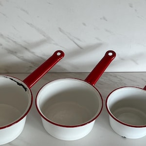 May include: Three white enamel saucepans with red handles and rims. The pans are of varying sizes, with a classic design. The handles have a hole for hanging. The pans are arranged in a row, showcasing their different sizes.