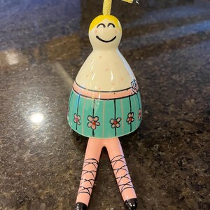 May include: A ceramic ballerina figurine with a smiling face, a white body with pink spots, and a turquoise skirt decorated with pink flowers. The figurine has pink legs with black crisscross ribbons.