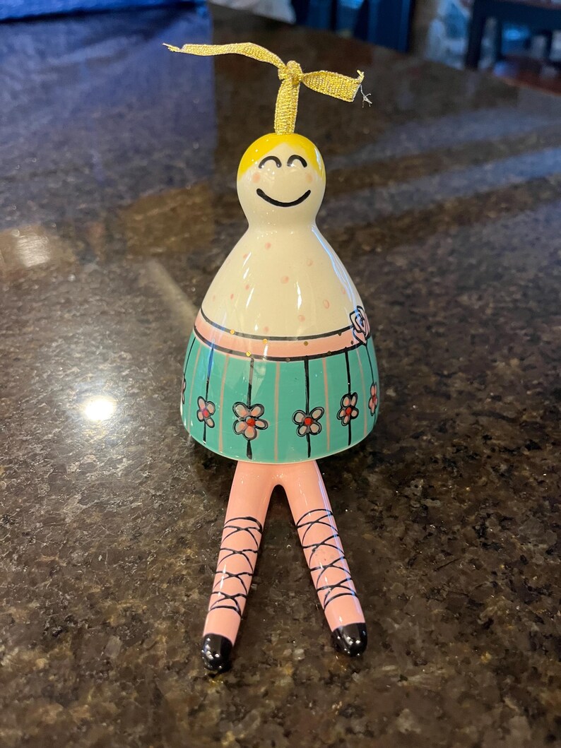 May include: A ceramic ballerina figurine with a smiling face, a white body with pink spots, and a turquoise skirt decorated with pink flowers. The figurine has pink legs with black crisscross ribbons.
