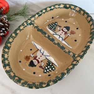 May include: Oval, divided serving dish with a tan interior and a green, star-patterned border. The dish features two snowmen with skis and a Christmas tree design. Red ornaments and pine needles are in the background.