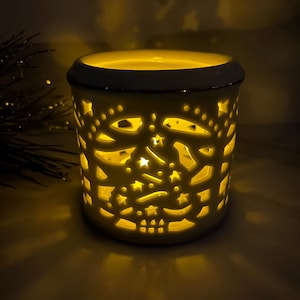 May include: White ceramic candle warmer with a cut-out design of stars and geometric shapes. The warmer is lit and glowing yellow.