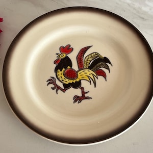 May include: A round ceramic plate with a rooster design. The plate has a cream-colored center with a brown rim. The rooster is depicted in black, yellow, red, and brown.