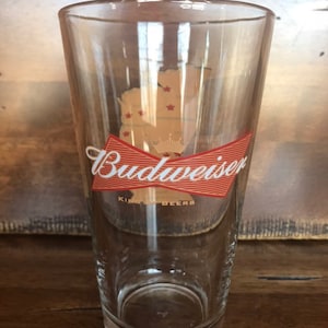 May include: Clear pint glass with the Budweiser logo in red and white. The logo includes a bow tie design and the words "Budweiser" and "King of Beers". A gold-colored outline of a state with red stars is also visible.