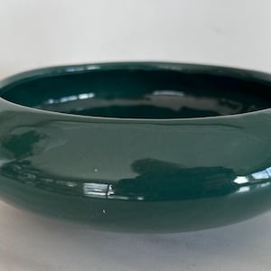 May include: A glossy, dark teal ceramic bowl with a wide, shallow design. The bowl reflects light, and is next to a cluster of orange and yellow flowers. A decorative piece for the home.