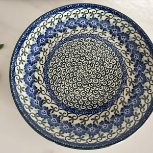 May include: A round ceramic plate featuring a central pattern of dark blue swirling lines surrounded by a border of blue flowers with green leaves on a cream-coloured background. The plate has a dark blue rim and a smooth, glazed finish.