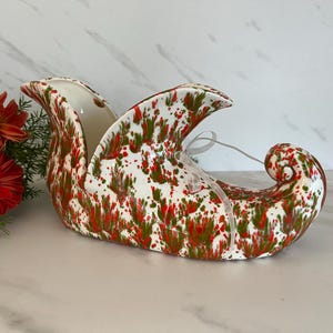 May include: A white ceramic Christmas stocking with a red and green splatter paint design. The stocking has a white ribbon tied around the top.