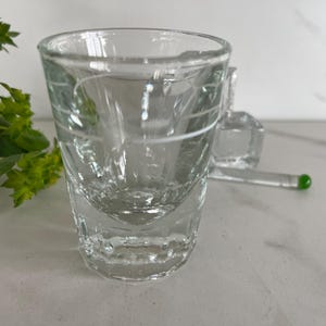 May include: A clear, textured shot glass with a thick base and a slightly flared rim. The glass has etched horizontal lines and is accompanied by a clear glass stirrer with a green tip. The shot glass is on a white surface.