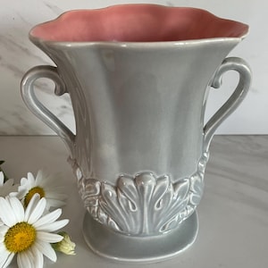 May include: A glossy gray ceramic vase with a scalloped rim and a soft pink interior. The vase features two curved side handles and a raised, embossed leaf-like pattern around the base. The exterior surface is smooth and reflective.