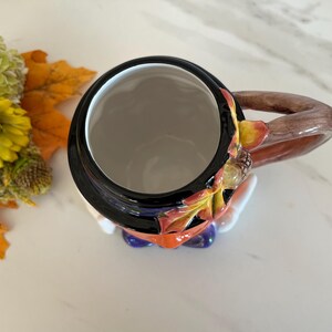 May include: A ceramic mug with a black rim and a brown handle. The mug is decorated with autumn leaves and acorns. The interior is white. The mug is a fall-themed item.