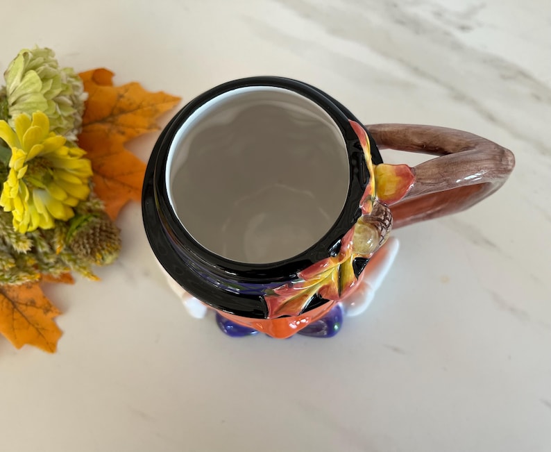 May include: A ceramic mug with a black rim and a brown handle. The mug is decorated with autumn leaves and acorns. The interior is white. The mug is a fall-themed item.