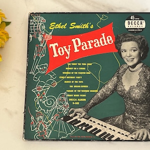 May include: Vintage vinyl record album cover for "Ethel Smith's Toy Parade." The cover features a smiling woman in a lace dress playing a piano. The album title is in a red banner, with a green background and illustrations.